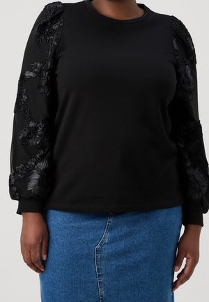 Personne portant un haut noir à manches longues avec des manches texturées florales et une jupe en jean bleue sur un fond neutre.