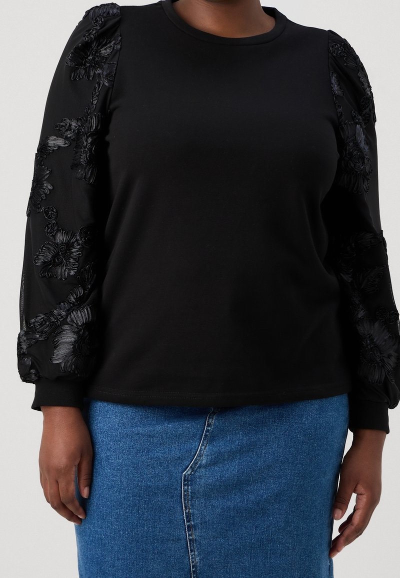 Personne portant un haut noir à manches longues avec des manches texturées florales et une jupe en jean bleue sur un fond neutre.