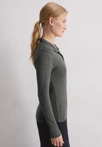 Kobieta w profilu, ubrana w dopasowany, długi rękaw, prążkowany szary sweter i czarne spodnie, stojąca przy gładkiej, jasnej ścianie.