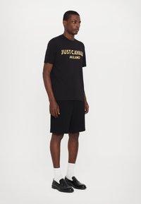 Černé bavlněné tričko s nápisem "JUST CAVALLI MILANO" v zlaté barvě, spárované s černými šortkami a černými mokasíny, doplněné bílými ponožkami.