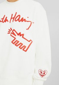 Vit sweatshirts med djärv röd grafisk tryck av en handgest på framsidan och ett hjärta med ansikte på ärmen. Mjuk textur.