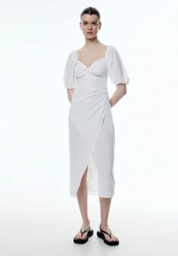 Weißes, knielanges Kleid mit enganliegendem Oberteil, Puffärmeln und gedrehtem Frontdetail. Verfügt über einen seitlichen Schlitz und ein strukturiertes, leichtes Material.