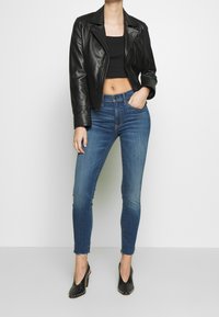 Chaqueta de cuero negra corta, top corto negro ajustado, jeans ajustados azules con dobladillo deshilachado y zapatos de tacón negro con acentos de tachuelas.
