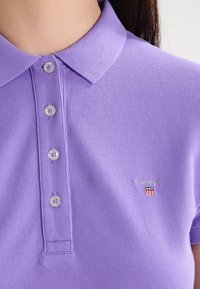 Fialové polo tričko s texturovanou látkou, má límec, pět knoflíků a mal�é vyšívané logo na levé hrudi.