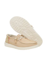 Scarpe in tessuto beige, texture con lacci bianchi, dettagli a nappina e suola decorata. Presentano un design morbido e flessibile con forma arrotondata.