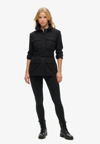 Chaqueta de mezclilla negra con cintura ceñida por cinturón, cierre de botones, dos bolsillos en el pecho y mangas enrolladas, combinada con jeans skinny negros y botas de combate.