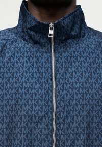 Κοντινό πλάνο ενός ναυτικού μπλε μπουφάν με επαναλαμβανόμενο μοτίβο "MK" και ένα ασημί φερμουάρ μερικώς κλεισμένο.