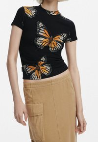 Top negro acanalado con estampado de mariposas en naranja y blanco, cuello redondo y mangas cortas. Combinado con pantalones estilo cargo en beige.