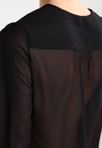 Blouse noire avec un dos transparent, une texture douce, un col rond et des plis discrets le long du dos pour plus de détails.