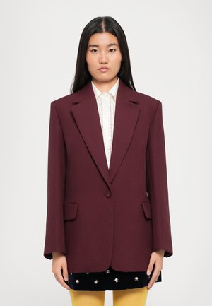 Burgundy oversized blazer with notched lapels, single button, and flap pockets, worn over a white textured top and black skirt.