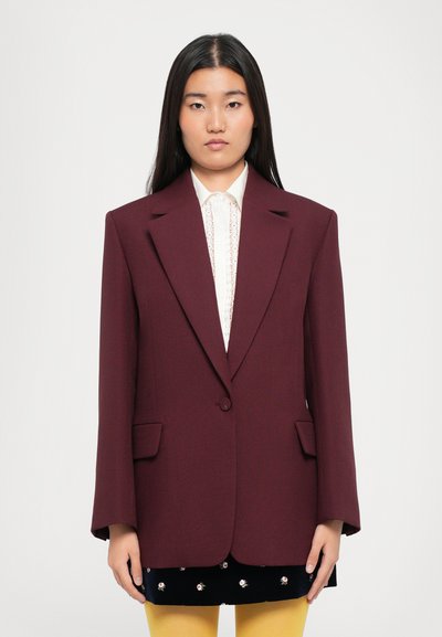 Burgundy blazer with structured shoulders, single button closure, and two front pockets, worn over a white shirt and layered with a dark skirt.