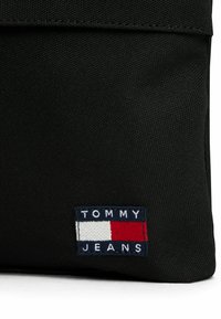 Borsa in tessuto nero con una patch logo di Tommy Jeans, decorata con accenti rossi, bianchi e blu. Il materiale presenta una finitura texture.