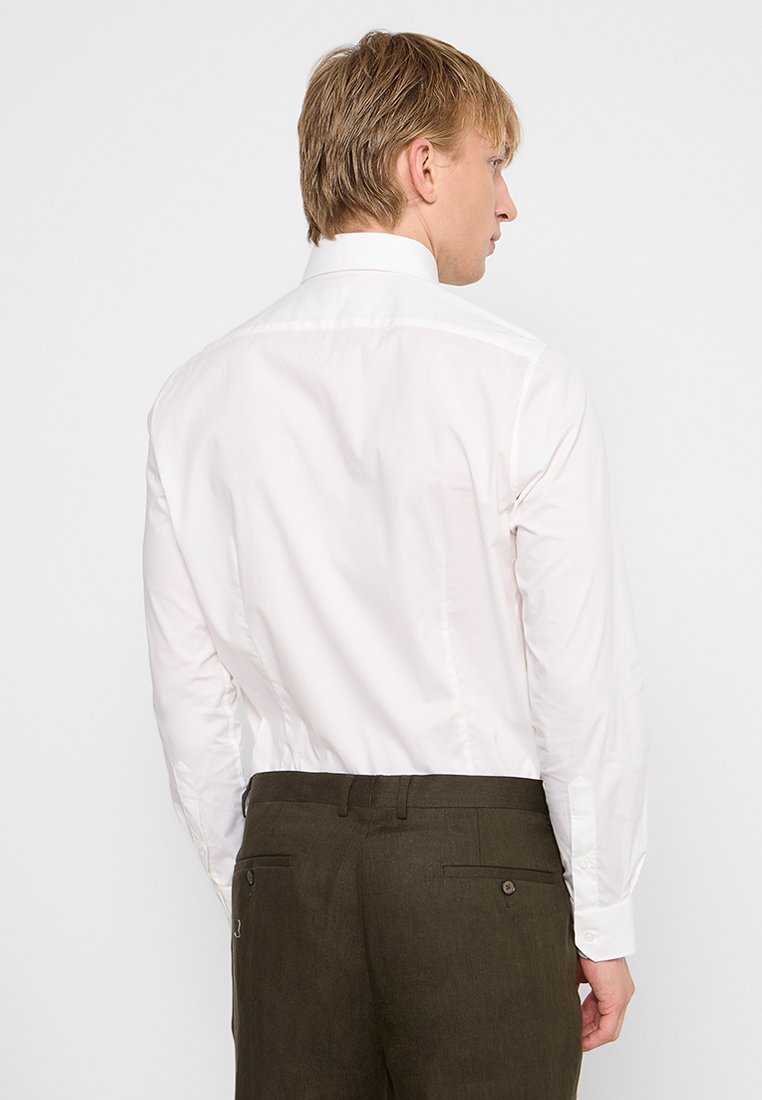 Blonde man die met zijn rug naar de camera staat, gekleed in een witte overhemd dat ingestopt is in donkerolijfgroene broek, tegen een effen achtergrond.