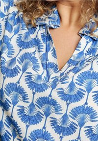 Camicia a fantasia floreale blu con grandi foglie bold su sfondo bianco. Presenta un colletto e un design a scollo a V. Tessuto morbido.