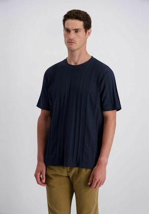 Man met kort donker haar draagt een marineblauw shirt met korte mouwen en mosterdkleurige broek, staande tegen een egale lichte achtergrond.