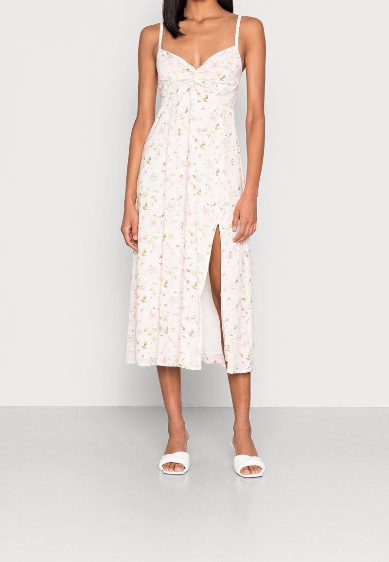 Vestido midi floral con un fondo rosa claro y acentos en verde y amarillo, un corpiño ajustado, tirantes finos y una abertura lateral.