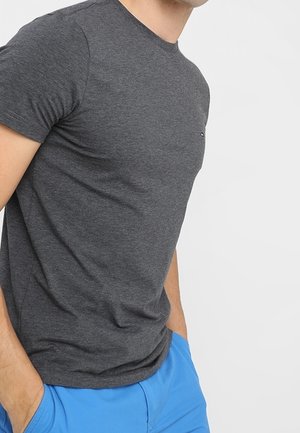 Mężczyzna ubrany w dopasowany, ciemnoszary t-shirt z krótkim rękawem i jaskrawo niebieskie spodnie, z rękami w kieszeniach, na jednolitym białym tle.
