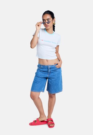 Femme portant un tee-shirt court blanc avec le texte "surfer boys", short en denim longueur genou, sandales rouges, ajustant de petites lunettes de soleil, avec des cheveux tressés.
