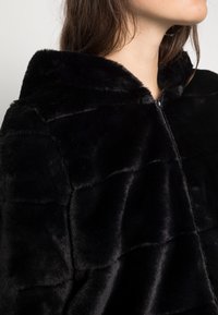 Giacca in pelliccia sintetica nera con chiusura a zip, dotata di cappuccio e motivo trapuntato per conferire texture. Lucentezza morbida e superficie soffice.