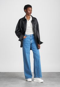 Jachetă din piele neagră, oversize, cămașă polo cu dungi albe, blugi evazați albaștri și adidași cu platformă albi. Textură netedă și croială lejeră.