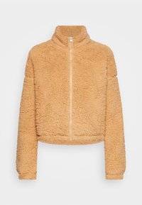 Veste zippée courte en tissu beige doux et texturé, avec un col haut et des manches longues. Dotée d'une fermeture éclair centrale.