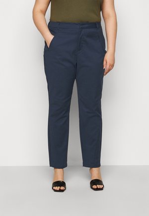 Kvinde iført marineblå tapered bukser med lommer, olivengrøn top og sorte åbne hælede sandaler, stående mod en ensfarvet baggrund.