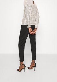 Paillettenbesetztes schulterfreies Top in Silber über transparentem Stoff, kombiniert mit schwarzen, schmal zulaufenden Hosen und schwarzen, römischen High-Heel-Sandalen mit Riemchen.