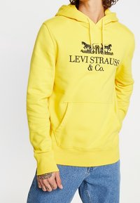 Sudadera con capucha amarilla y bolsillo frontal. Presenta el logotipo de Levi Strauss & Co. en negro y dos gráficos de caballos. Material de mezcla de algodón.