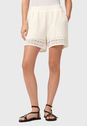 Femme portant un short en crochet couleur crème avec un ourlet festonné et des sandales plates noires à lanières, debout devant un fond clair.