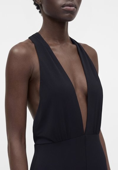 Robe noire sans manches avec un profond décolleté en V portée par une personne à la peau foncée, montrant les épaules et le haut de la poitrine sur un fond uni.