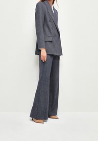 Traje en espiga azul marino con una chaqueta cruzada y pantalones de pierna ancha. Textura suave y sutil brillo; combinado con tacones de punta beige.