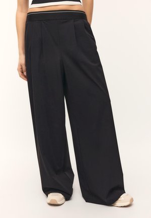 Žena v voľných čiernych širokých nohaviciach s bočnými vreckami a béžovými teniskami, stojaca pred jednoduchým svetlým pozadím.