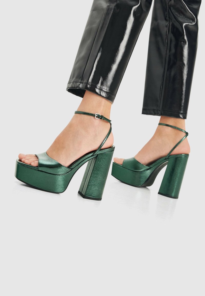 Pieds portant des sandales à talons hauts plateforme vert brillant avec brides à la cheville et pantalon court noir brillant sur fond blanc.