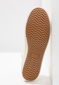 Semelle d'une chaussure TOMS de couleur claire montrant un motif de bande de roulement en zigzag et un logo rectangulaire sur une semelle extérieure en caoutchouc brun.