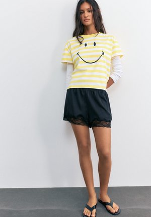 Jeune femme debout contre un mur blanc, portant un t-shirt rayé jaune avec un smiley, un short noir avec bord en dentelle et des sandales noires.