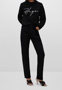 Schwarzer Pullover mit weißem, geschwungenem "Hugo"-Logo, kombiniert mit maßgeschneiderten schwarzen Hosen. Das Model trägt schwarze Absatzsandalen.