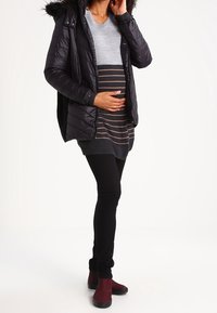 Czarna kurtka puchowa z kapturem wyłożonym futrem; szary sweter w paski; czarne legginsy; burgundowe botki do kostki. Skupienie na komforcie i fasonie odzieży ciążowej.