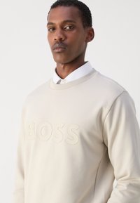 Sudadera beige con cuello redondo, que presenta un logo en relieve de "BOSS" en el centro. Tejido suave con una textura lisa y un diseño minimalista.