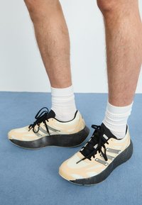 Des chaussures de sport beiges et noires sont dotées de panneaux en maille, d'une semelle noire épaisse et de lacets. Elles sont portées avec des chaussettes blanches et se tiennent sur une surface bleue.