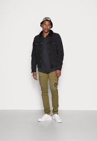 Black denim jacket with shearling collar, black t-shirt, olive cargo pants with patch, and white sneakers. Grey bucket hat with pattern.