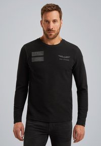 Schwarzes Langarm-T-Shirt aus Baumwolle mit Rundhalsausschnitt, das geprägte Patches und Details auf der Brust und dem Ärmel für zusätzliche Textur aufweist.