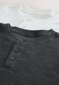 Dve pletené henley košele so vzorom vaflí, jedna čierna a jedna biela, každá s okrúhlym výstrihom a prednými gombíkmi, rozložené na rovnej ploche.