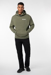 Man wearing an olive green hoodie with "WORLDWIDE" text, black cargo pants, and black sneakers, standing with hands in hoodie pocket.