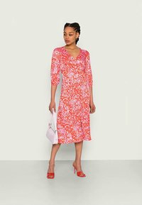 Vestido em padrão vermelho e rosa com mangas três quartos, decote em v e comprimento até ao joelho. Acompanhado de uma mala branca e sapatos de salto vermelhos.