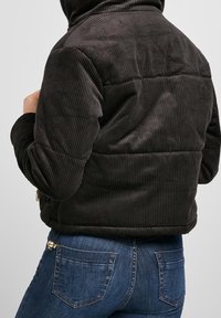 Kurtka sztruksowa w ciemnym brązie, zapinana na zamek, o prążkowanej teksturze. Połączona z niebieskimi dżinsami, z detalami kieszeni na plecach i subtelnym przeszyciem.