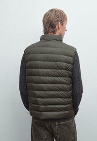 Ung mand med kort blond hår iført en olivengrøn quiltet vest over en mørkegrå langærmet skjorte, vendt væk mod en ensfarvet baggrund.