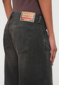 Μαύρο τζιν από τζιν ύφασμα με εμφανές λογότυπο σε πορτοκαλί και μπλε, με αντιθετικές ραφές και διαγώνιες τσέπες.