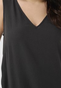 Femme portant un haut noir sans manches à col en V avec des cheveux foncés ondulés visibles sur le côté droit.