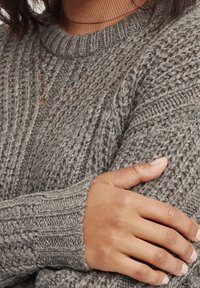 Osoba w grubym szarym swetrze z dzianiny z prążkowanym kołnierzem i mankietami, z ręką opartą na przeciwnym ramieniu, na tle brązowego kołnierza koszuli.
