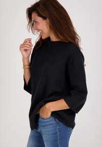 Vrouw met lang bruin haar die glimlacht, gekleed in een losse zwarte trui, gouden creolen, armbanden en blauwe spijkerbroek, hand in de zak.